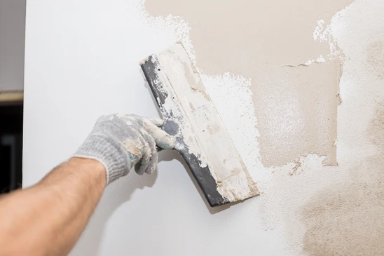 Intérieur de chantier avec ouvrier appliquant enduit sur mur. Main gantée tenant platoir métallique, lissage de pâte blanche sur surface murale. Travaux de rénovation en cours.