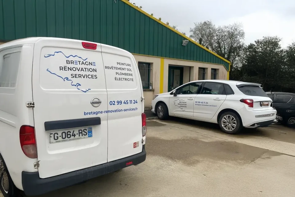 Extérieur d'entreprise de rénovation avec véhicules utilitaires blancs stationnés. Bâtiment industriel à bardage métallique vert et façade beige avec menuiseries vertes.