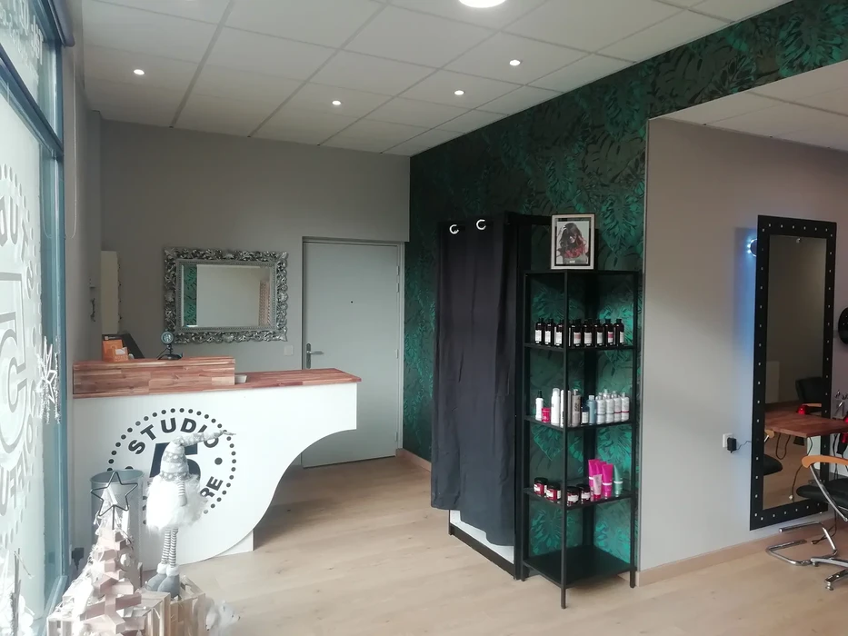 Intérieur de salon de coiffure moderne avec réception au comptoir en bois, miroir baroque argenté au mur gris, étagère noire avec produits capillaires sur mur vert texturé. Espace professionnel aménagé avec fauteuil de coiffure, carrelage beige au sol.