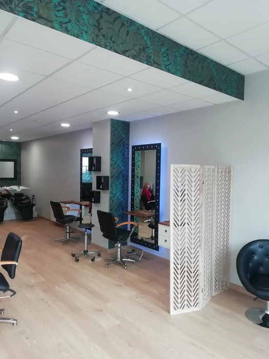 Salon de coiffure moderne avec plusieurs postes de travail équipés de fauteuils hydrauliques noirs et miroirs éclairés. Une cliente est installée pour une prestation. L'espace présente un plafond suspendu avec motifs végétaux turquoise, sol en parquet stratifié, cloison décorative blanche ajourée géométrique. Éclairage LED encastré, bac de lavage visible, mobilier professionnel noir et rangements muraux.