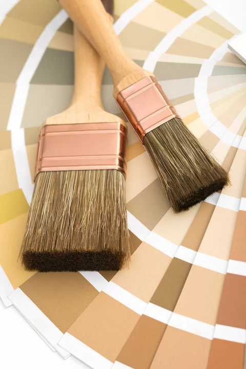 Atelier de peinture en bâtiment avec pinceau plat professionnel taille 60 posé sur établi en bois. Nuancier de couleurs ouvert présentant palette chromatique complète en arrière-plan.